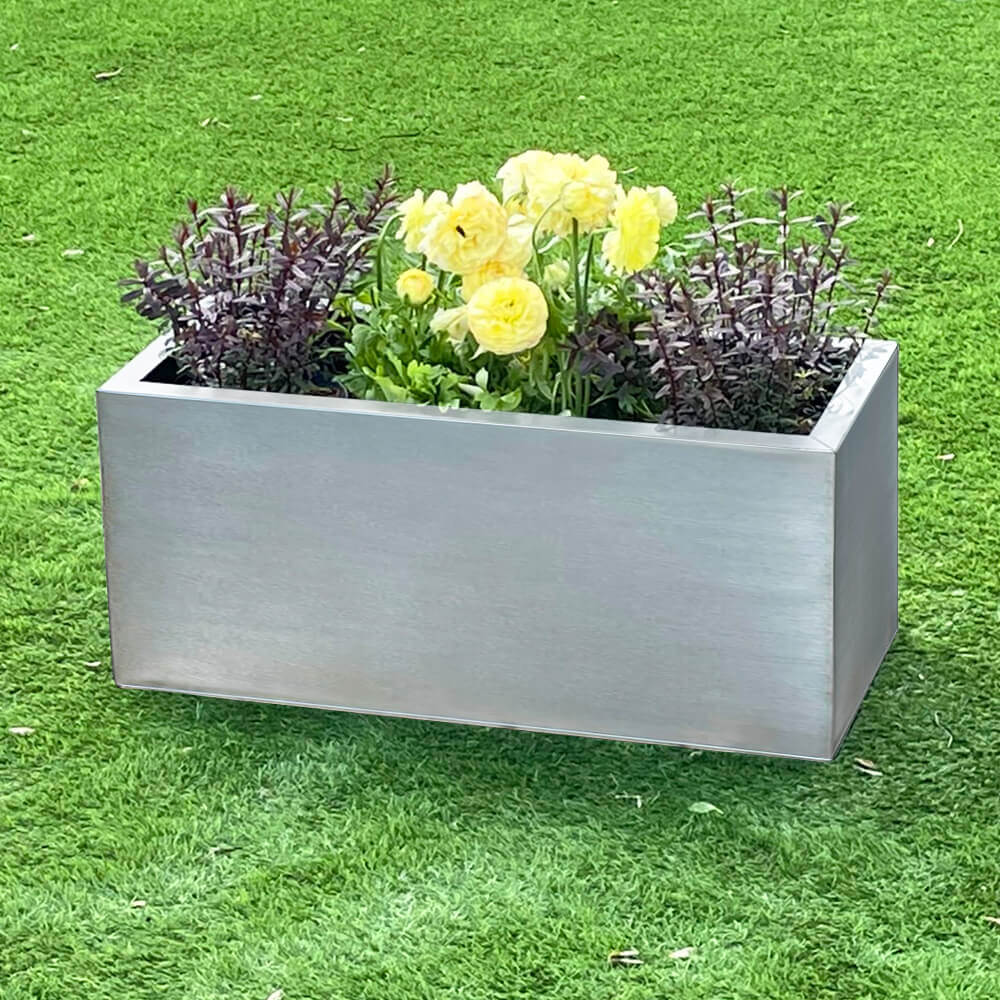 A silver zinc metal trough garden planter filled with various plants and flowers, placed on a green grass surface.