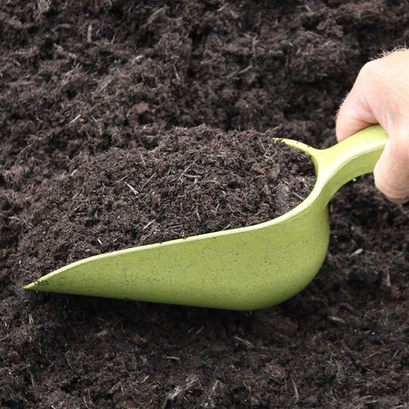 An olive green bamboo compost scoop filled with soil, held by a hand above a pile of soil.