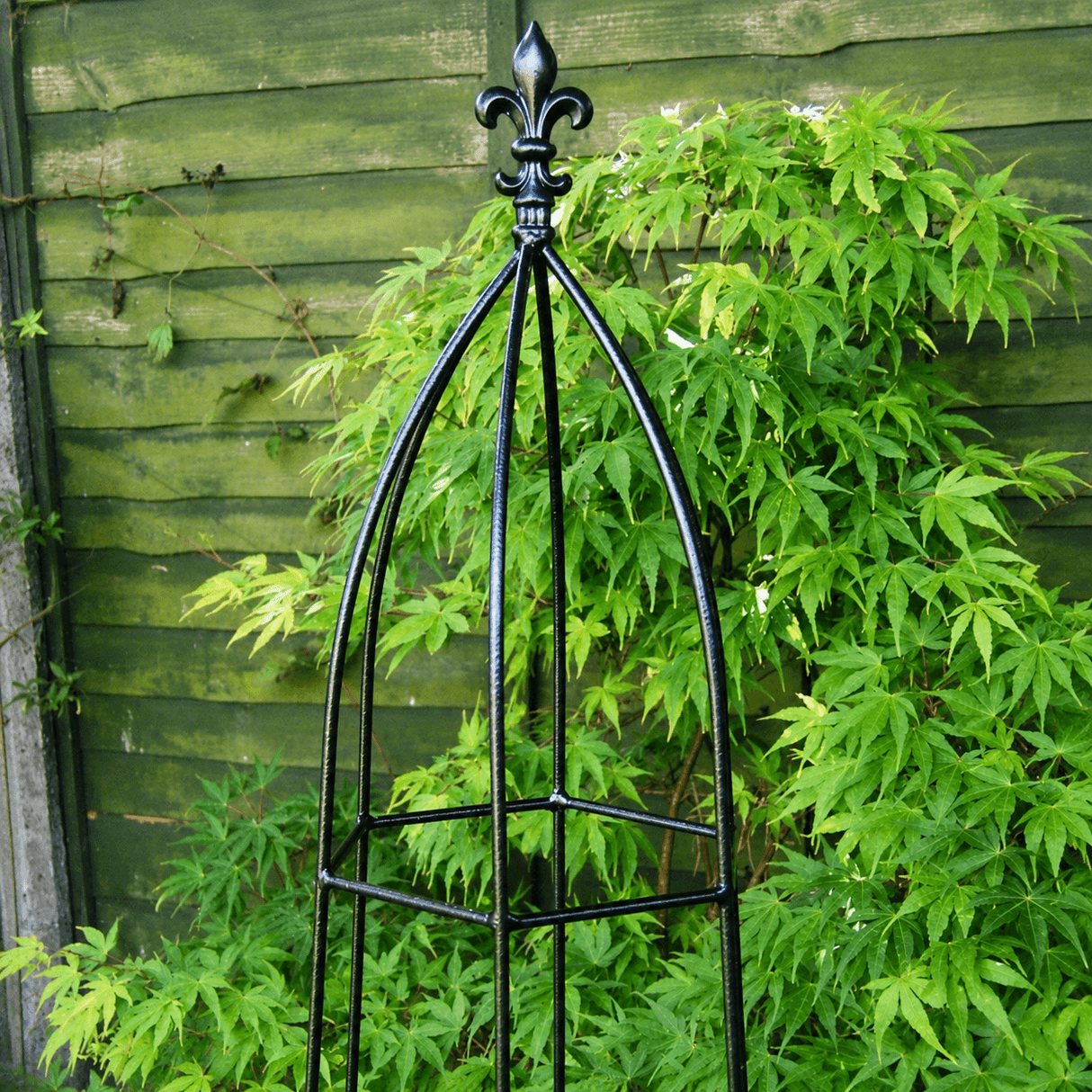 Black metal garden obelisk with decorative top against a green leafy background