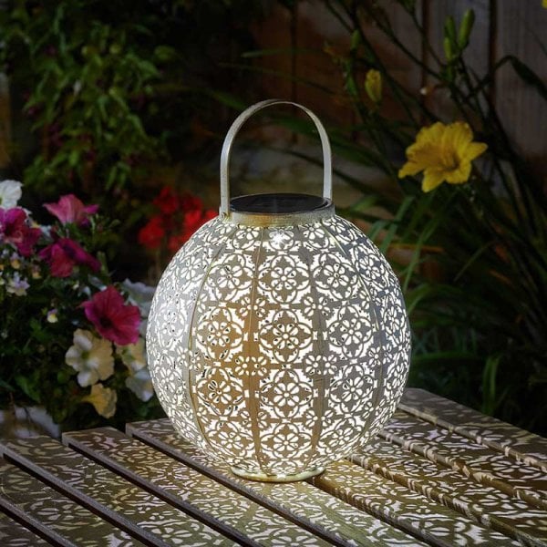 A cream-colored metal solar lantern with a geometric pattern, placed on a table in a garden setting, emitting a warm glow.