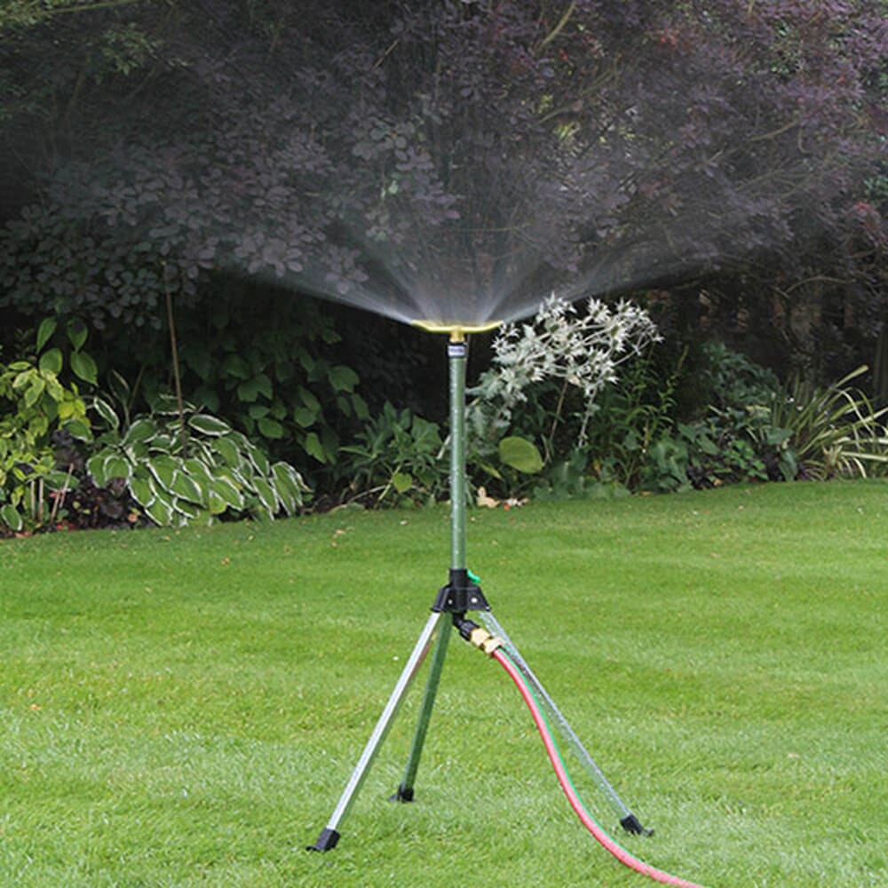 Garden sprinkler on a stand with a hose, spraying water on a grassy area with plants in the background.