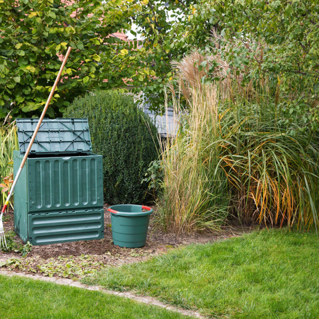 A green Eco-King composter located in a garden setting, with a green bucket next to it and foliage in the background.