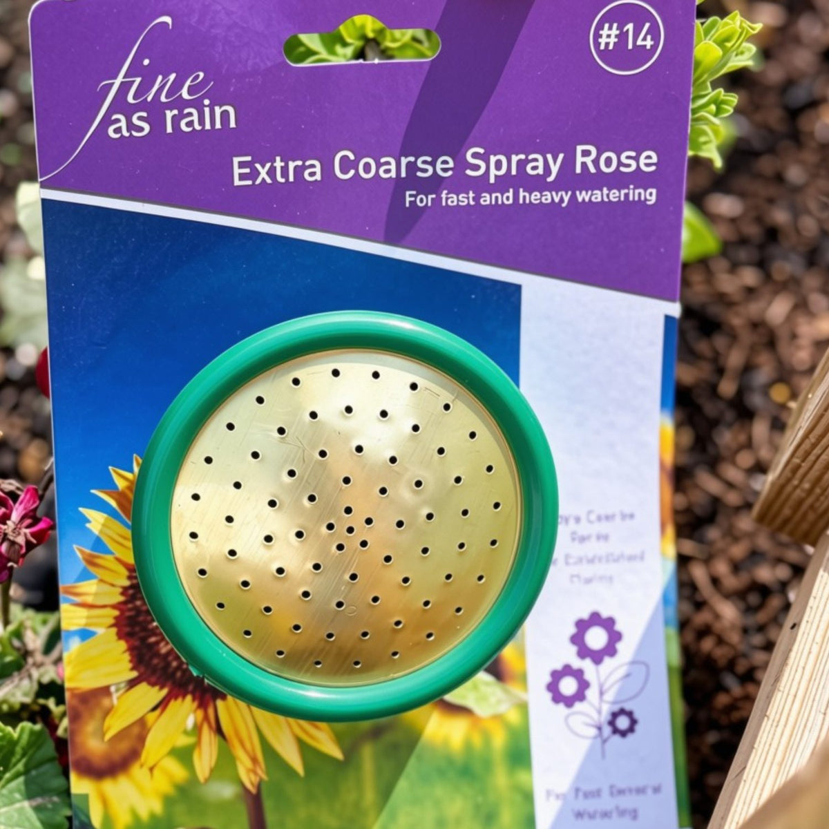 Fine as Rain Extra Coarse Spray Rose garden watering can with a green rim and gold spray head, attached to its purple and blue retail packaging card, photographed outdoors near plants and mulch.