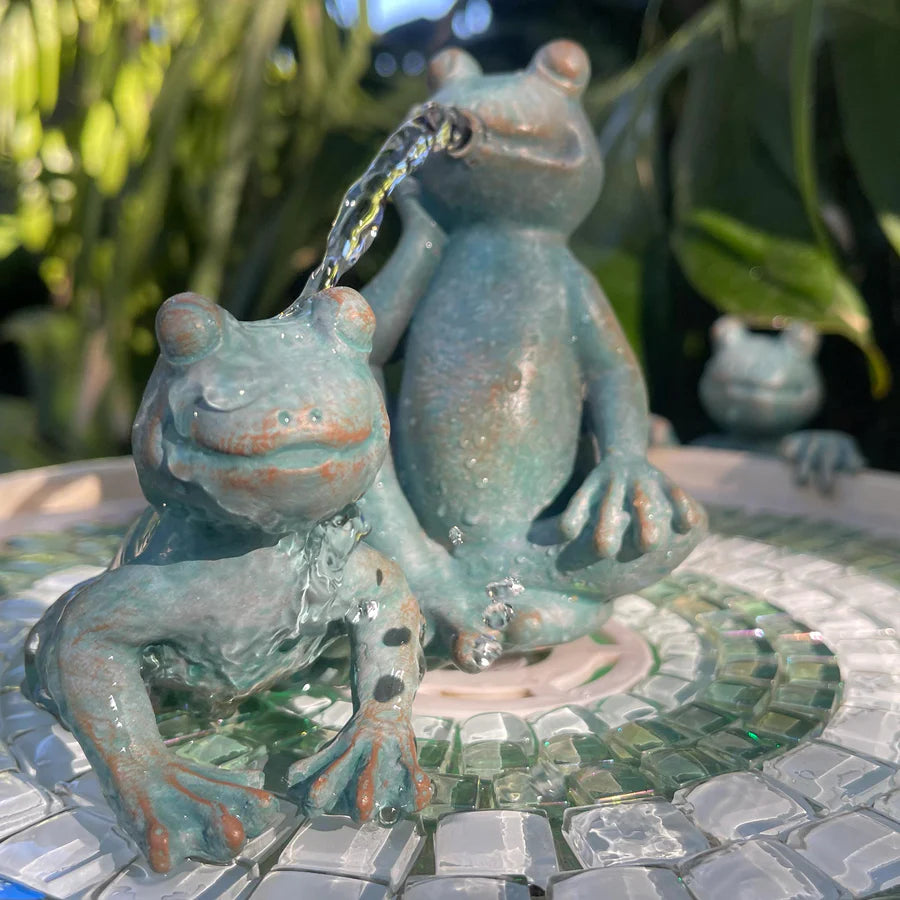 Frog sculpture pouring water from a fountain into another frog's mouth on a mosaic stone surface.