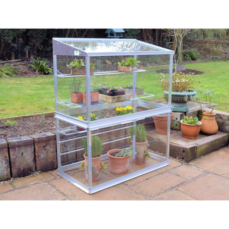 A small aluminum-framed greenhouse with transparent glass panels and multiple shelves, containing potted plants and labeled as 'Mini Greenhouse Growhouses'.
