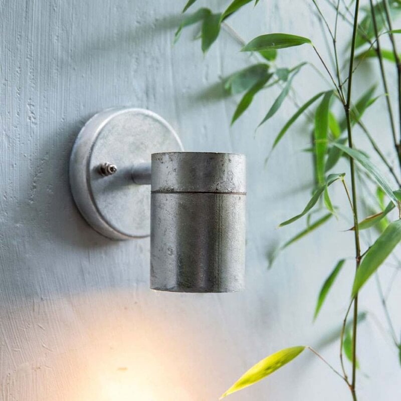 A silver galvanised steel exterior wall light mounted on a wall, with a cylindrical shape and a beam of light emitting from it, set against a natural background with foliage.