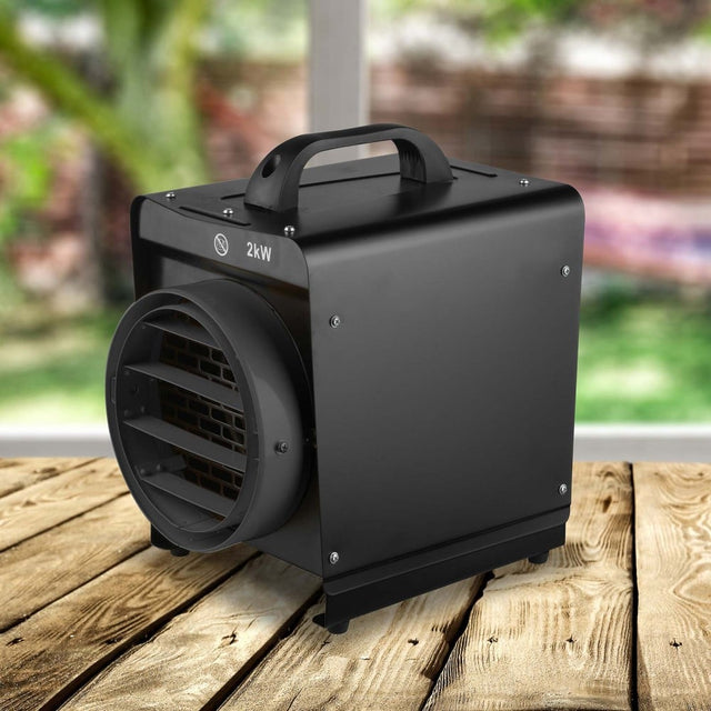 A black 2kW electric greenhouse heater with a handle on top, situated on a wooden surface with a blurred background of a greenhouse.