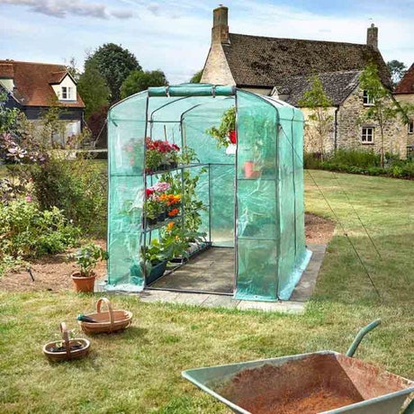 A small free-standing greenhouse with a green PE cover and white steel frame, set in a garden with various plants inside, a wheelbarrow, and a house in the background.