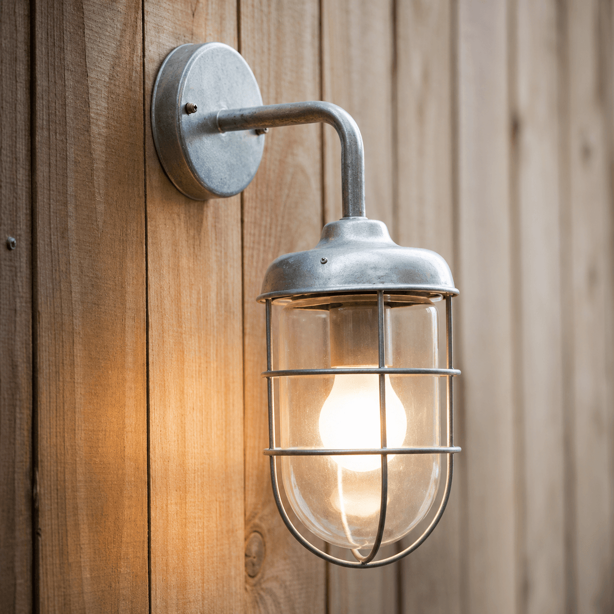 A wall-mounted garden light with a galvanised steel cage and a lit bulb, attached to a wooden surface.