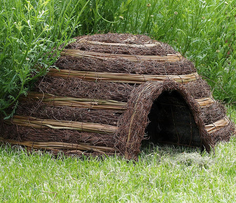 A rustic, igloo-shaped hedgehog house made of straw and branches, designed to resemble a natural den.