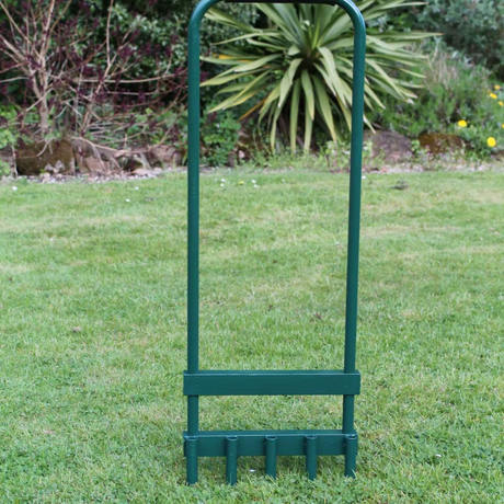 A green hollow tine lawn aerator with a foam support handle, standing on grass with a shrub in the background.