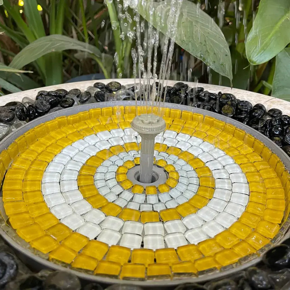 Mosaic fountain with yellow and white tiles and black stones, surrounded by green foliage.