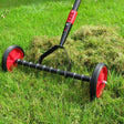 A red and black push along lawn scarifier with a metal frame and two wheels, being used on a lawn.
