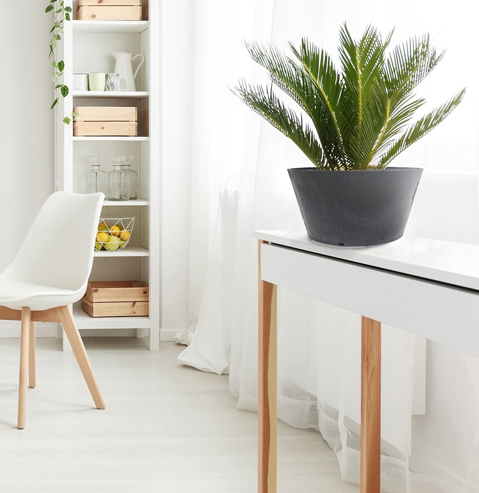 A slate-colored Octavia garden planter made from recycled rubber, placed on the floor next to a wooden cabinet and a decorative vase, containing a green plant.