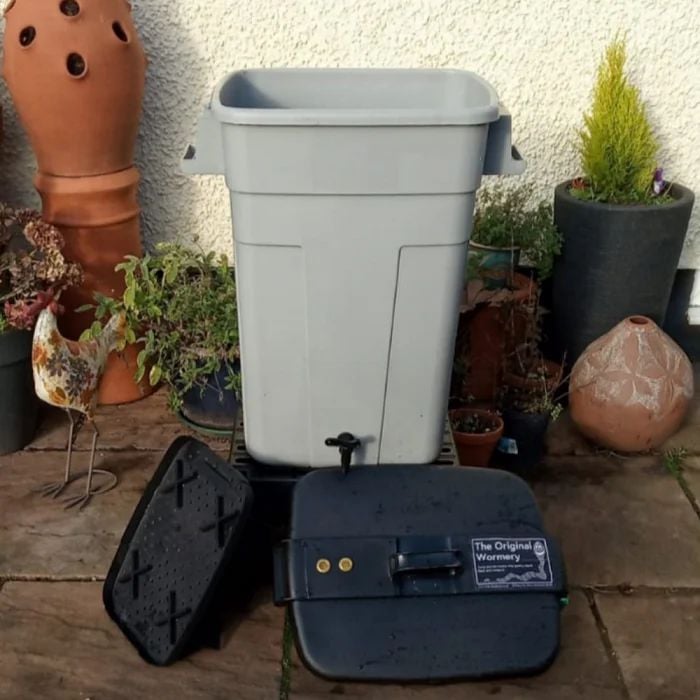 A grey Deluxe Original Wormery bin with stand, surrounded by various garden ornaments and plants.