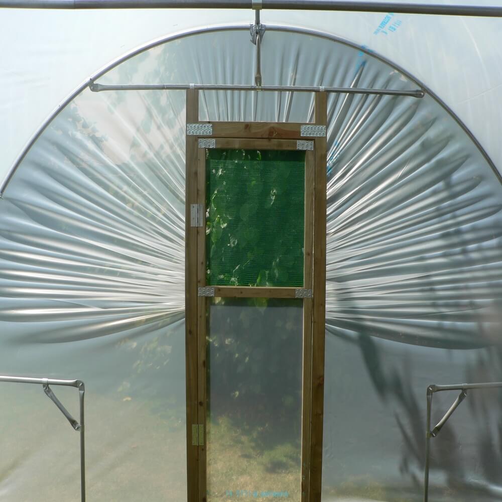 Clear plastic hoop house with a wooden door, showing greenery inside.