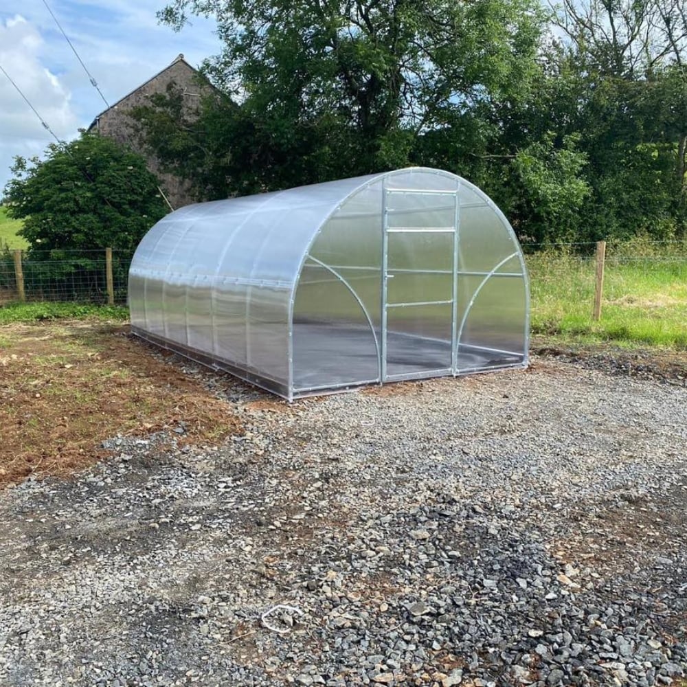 A polytunnel greenhouse with a metallic frame and clear polycarbonate covering, situated outdoors with a fence in the background.