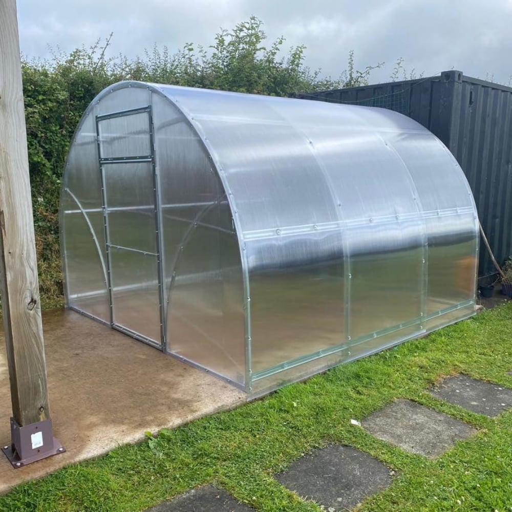 A polytunnel greenhouse with a metallic frame and clear polycarbonate covering, situated outdoors with a fence in the background.