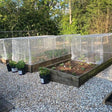A set of portable square cloche tunnels with a domed cage design, used to protect plants in a garden setting. The tunnels are set on a gravel surface and there are potted plants beside them.