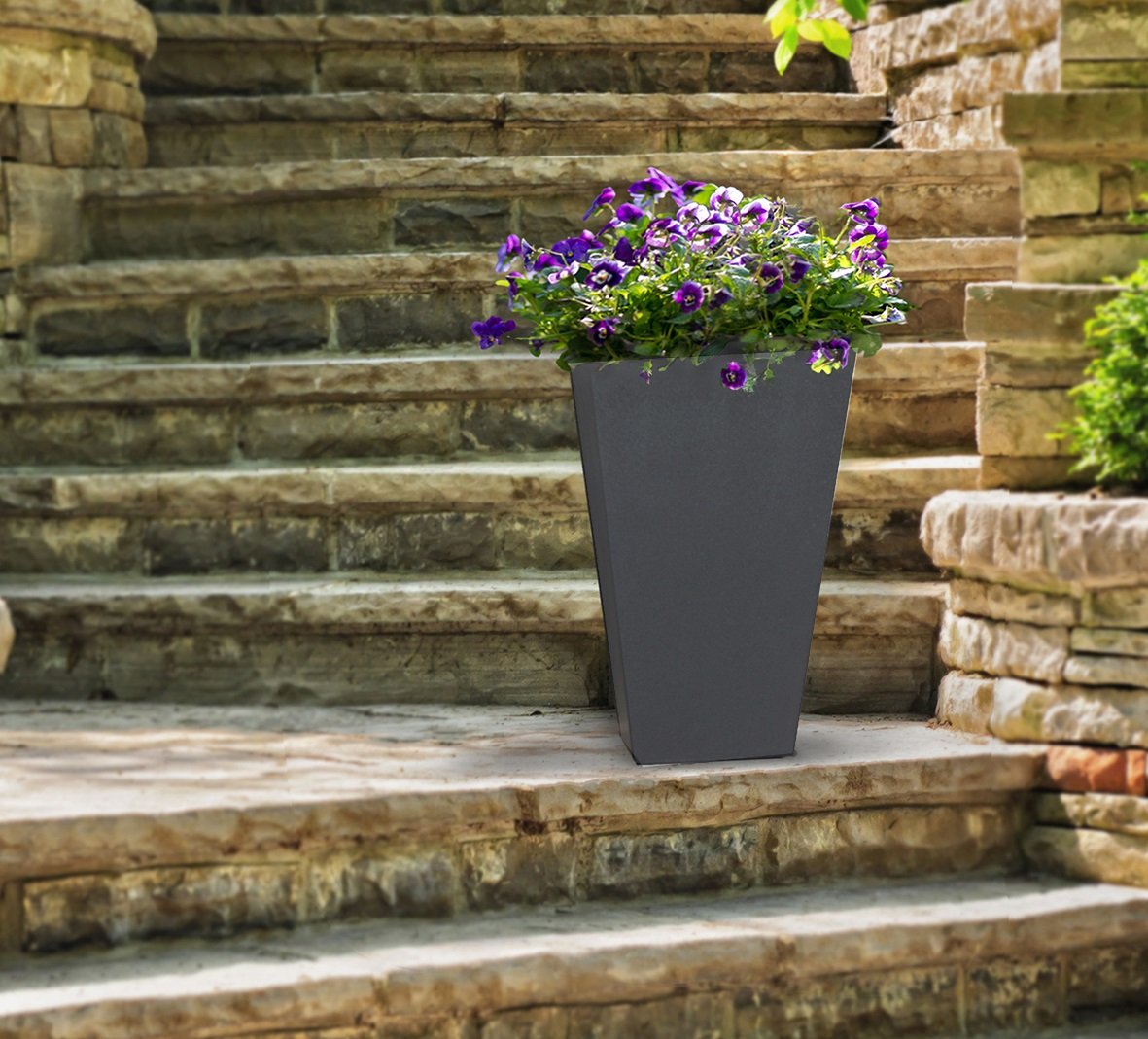 A gray, self-watering planter pot placed on a stone step, containing purple and green plants.