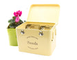 A vintage cream-colored metal seed storage tin with compartments, displayed alongside a pink potted plant.