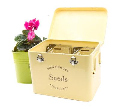 A vintage cream-colored metal seed storage tin with compartments, displayed alongside a pink potted plant.