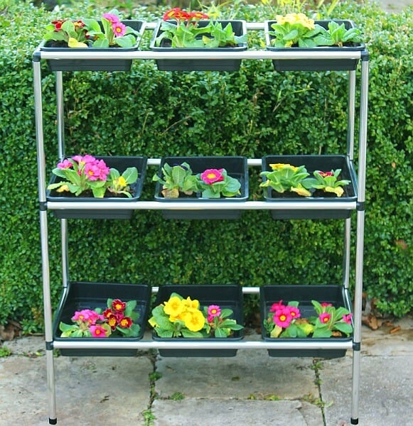 A three-tiered aluminum seed tray stand with various trays containing colorful plants, placed outdoors.