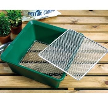 A green plastic garden sieve with a metal screen placed on top, situated on a wooden surface with plants in the background.
