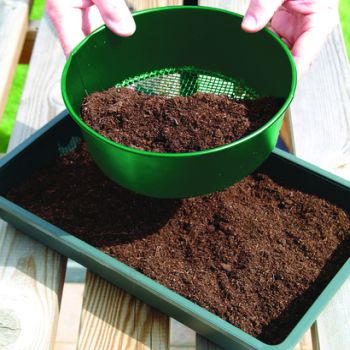 A green seed tray sieve with 3mm mesh being used to sieve soil into a tray.