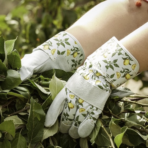 A pair of white gardening rigger gloves with green leaf patterns, worn by a person amidst green foliage.