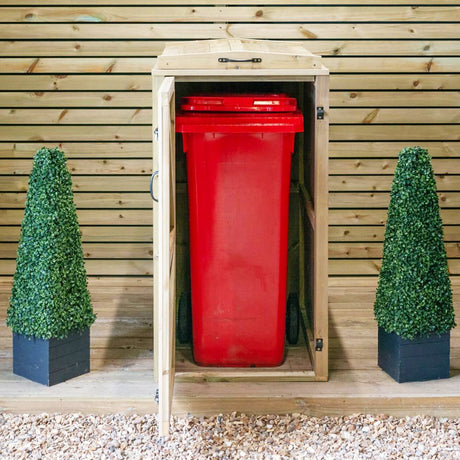 A single wooden wheelie bin storage unit with a red bin visible, placed on a gravel surface, flanked by two artificial green shrub pots.