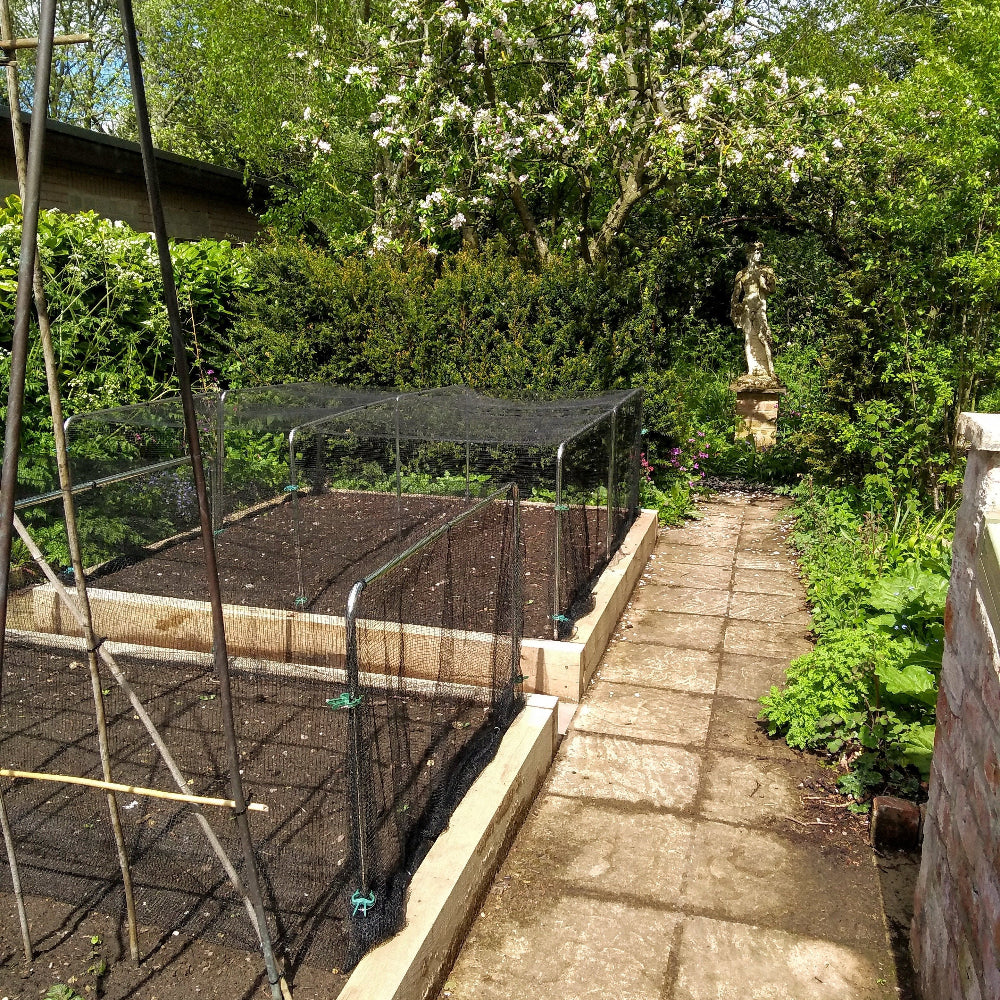 A garden tunnel kit with square aluminium hoops and a white netting covering, installed over young plants in soil.