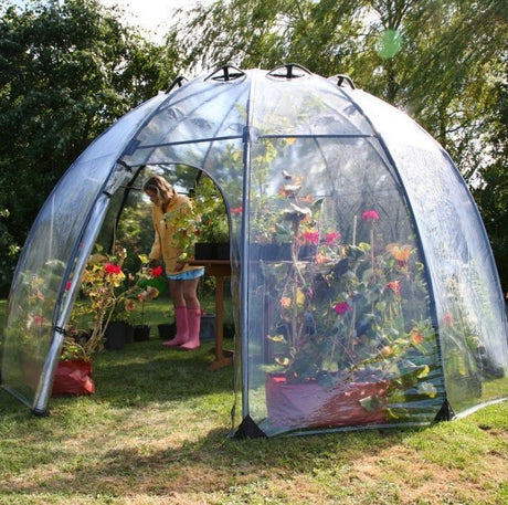 A large green dome greenhouse made of PVC with a fibreglass structure, set up outdoors with plants inside and a person standing outside.
