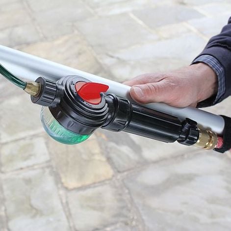 A hand holding a Swop Top soap dispenser with a hose, featuring a green liquid inside the dispenser and a red on/off dial on the top.