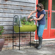 A person tending to plants inside a green, fabric-covered garden bed with a poly protection cover, placed on a metal frame outdoors.