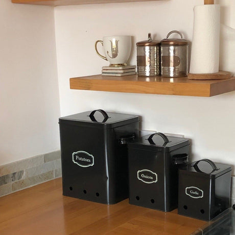 A set of three black kitchen storage tins with labels 'Potato', 'Onion', and 'Garlic' on them, placed on a kitchen counter.