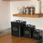 A set of three black kitchen storage tins with labels 'Potato', 'Onion', and 'Garlic' on them, placed on a kitchen counter.