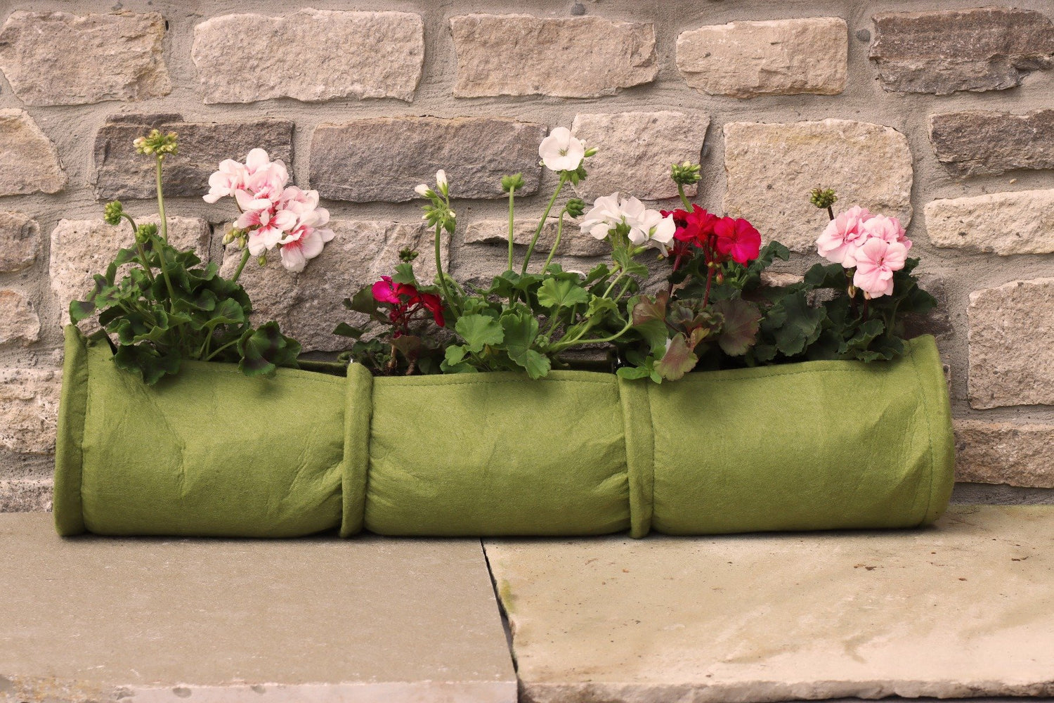 A green Vigoroot plant tube filled with soil and various flowers, placed on a stone surface against a brick wall.