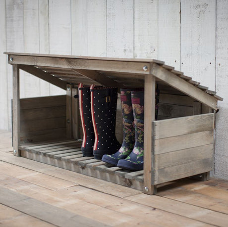 A wooden welly store with a slanted roof and 3 sides, containing up to 5 pairs of boots, placed on a wooden floor against a white wall.