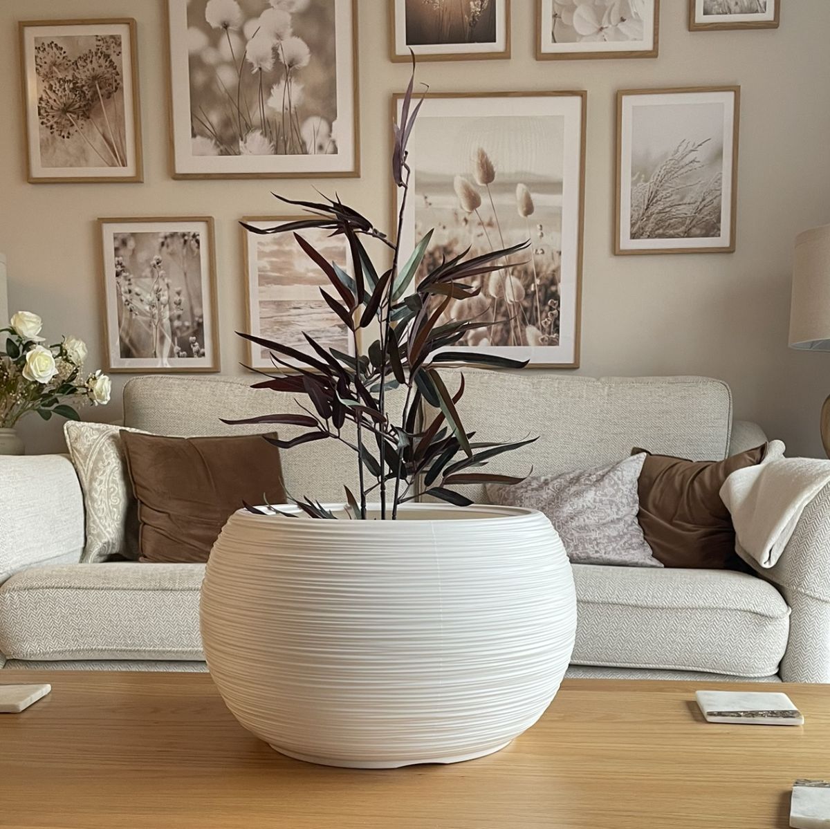 A round, white and grey textured plant pot with a green plant inside, placed on a wooden floor in front of a sofa with cushions and a picture wall in the background.