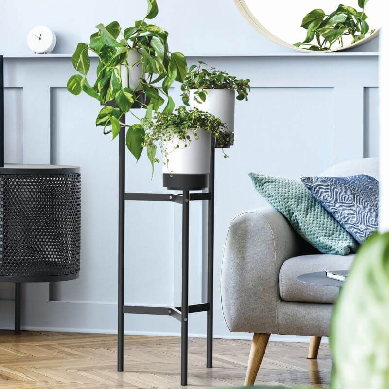 A white plant pot stand with three levels, featuring a sturdy industrial metal frame, placed in a room with various plants and a grey sofa.