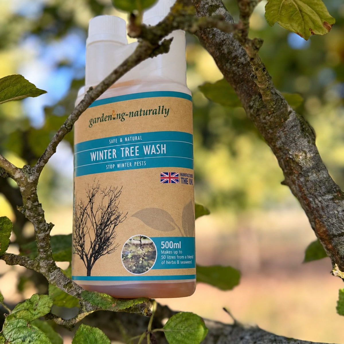 A 500ml bottle of gardenng-naturally Winter Tree Wash hanging from a tree branch outdoors, with green leaves and a blurred natural background.