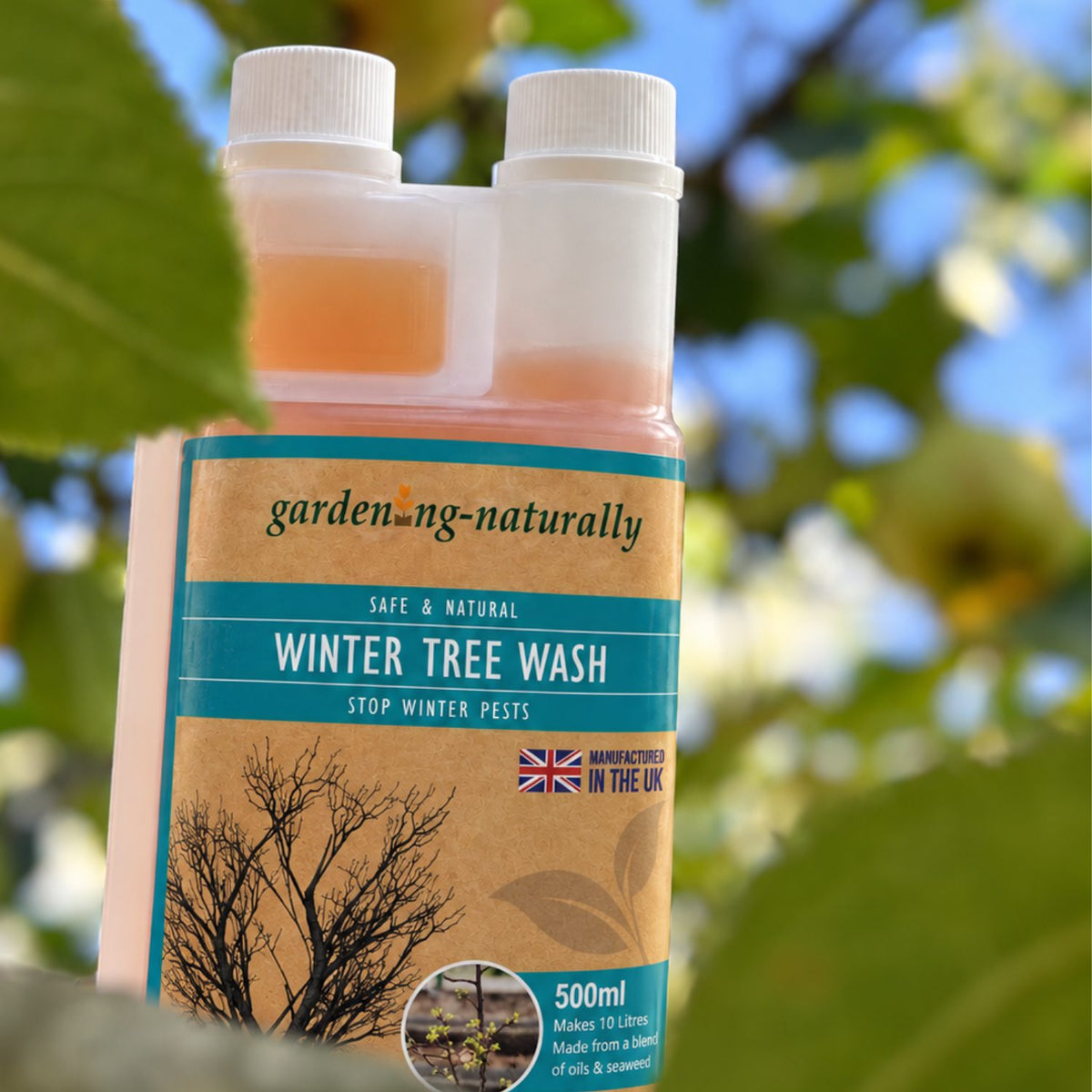 A bottle of gardening-naturally Winter Tree Wash with a brown kraft-style label and teal text, photographed outdoors with blurred green leaves and blue sky in the background.