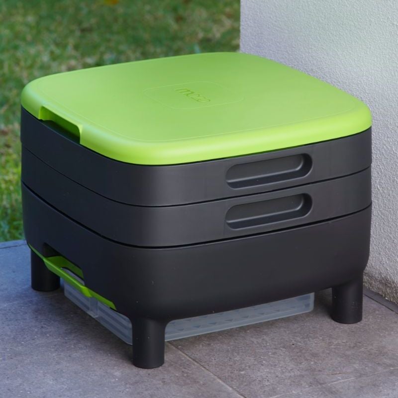 A black and green wormery system made of recycled material with two working trays, a lid, base, and a liquid collection tray, placed on a tiled surface outdoors.