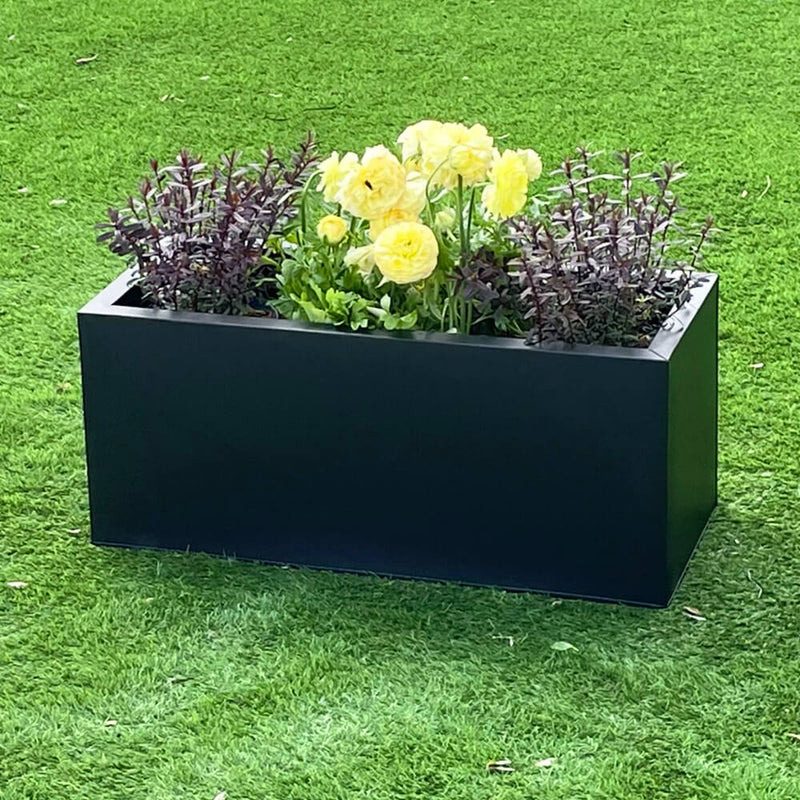 A black zinc metal trough garden planter filled with various plants and flowers, placed on a green grass surface.