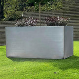 A large rectangular black zinc trough patio planter placed on a grassy area with a wooden fence in the background. The planter is empty and has a reflective surface.