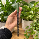 A hand holds a green and black laser level pen against a background of potted green plants.
