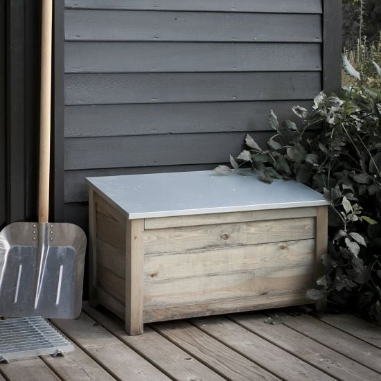 A rustic wooden outdoor storage box with a zinc hinged slated lid, placed on a wooden deck next to a wall and some plants, with a shovel leaning against the wall.