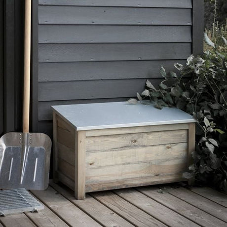 A rustic wooden outdoor storage box with a zinc hinged slated lid, placed on a wooden deck next to a wall and some plants, with a shovel leaning against the wall.