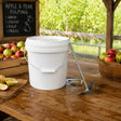 A white plastic scratting bucket with a metal handle sits on a wooden table in an apple orchard setting, with apples in crates